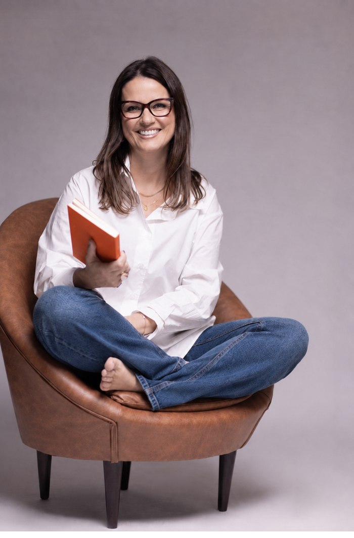 Andiara Neuwiem sorrindo, sentada em uma poltrona, com um livro nas mãos