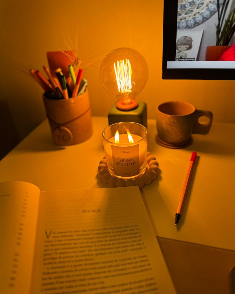 Mesa de estudo à noite, com vela, caderno e livro aberto, e a Andi na tela conduzindo um encontro online
